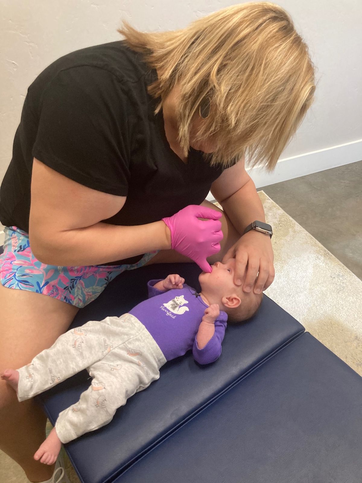A chiropractor wearing gloves gently works inside a baby's mouth as the baby lies on a padded table