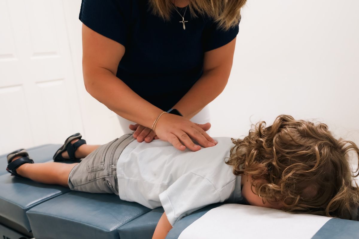 A chiropractor's hands gently adjusting the back of a curly-haired child lying face down on an adjusting table