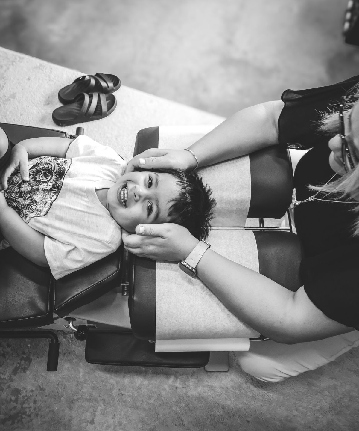 Black-and-white photo of a laughing boy lying on an adjusting table while a chiropractor gently holds his head