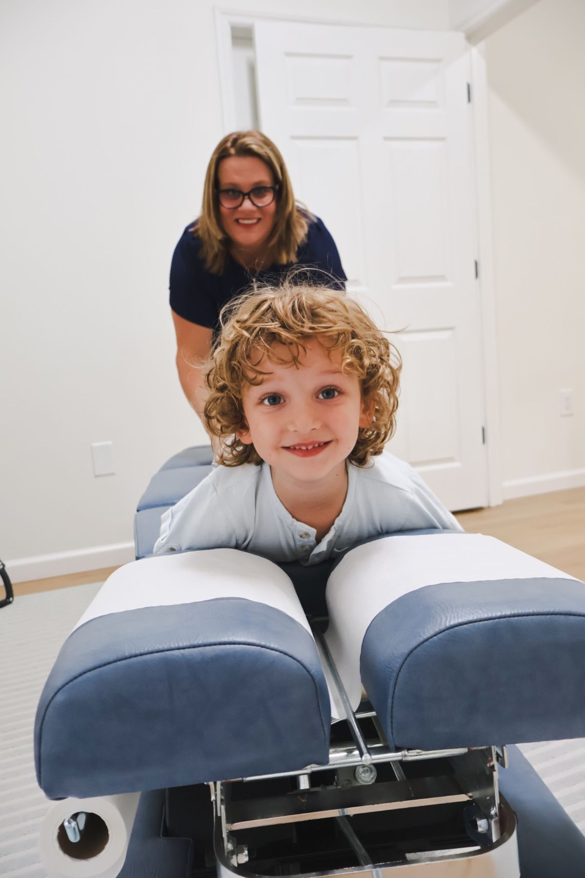 A smiling curly-haired boy at a chiropractic table, with a chiropractor standing behind him