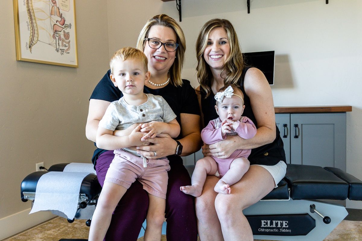 Two Vitality team members sitting on an adjusting table, each holding a young child