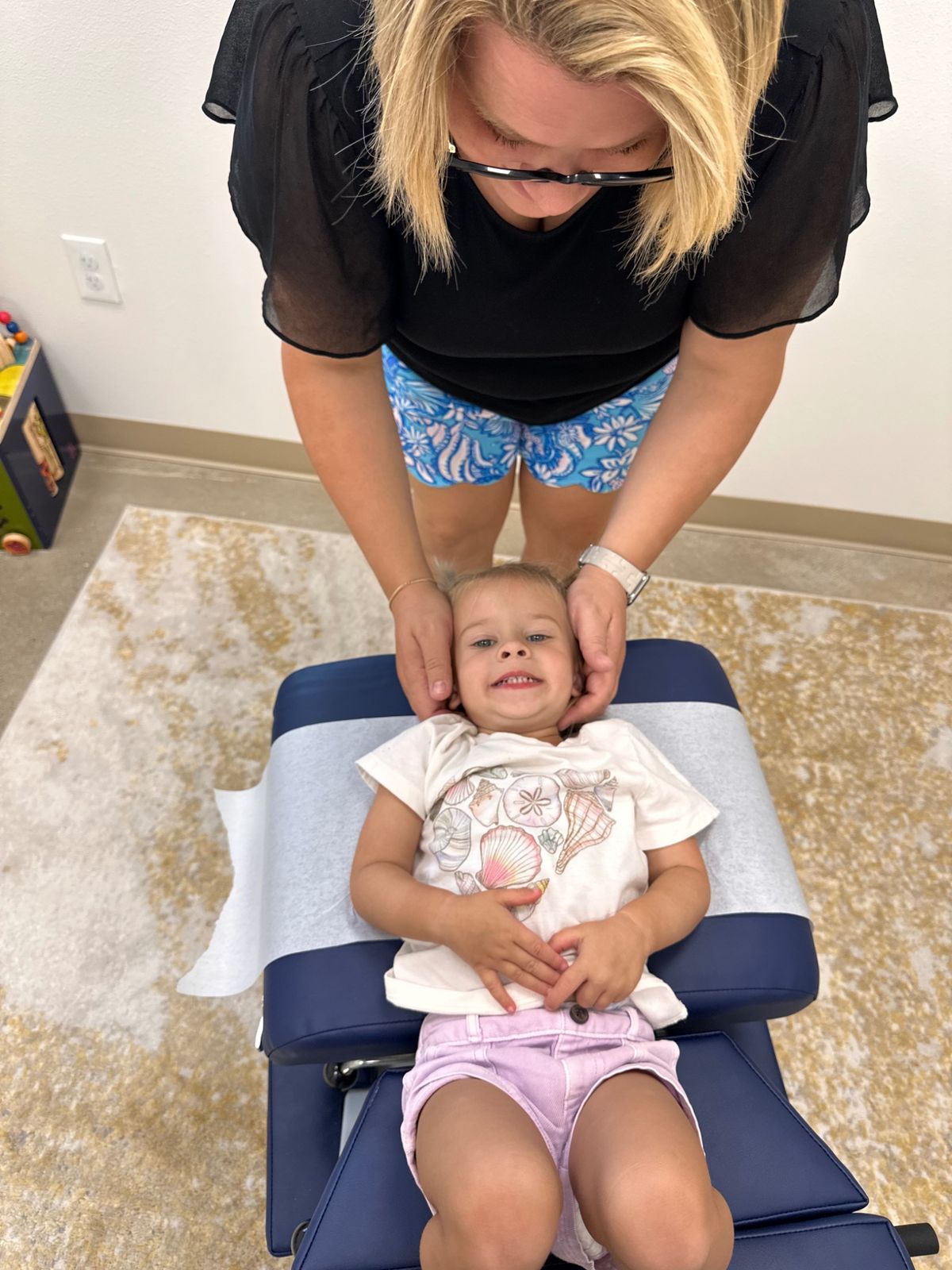 A smiling toddler lies on an adjusting table while a chiropractor gently cradles her head
