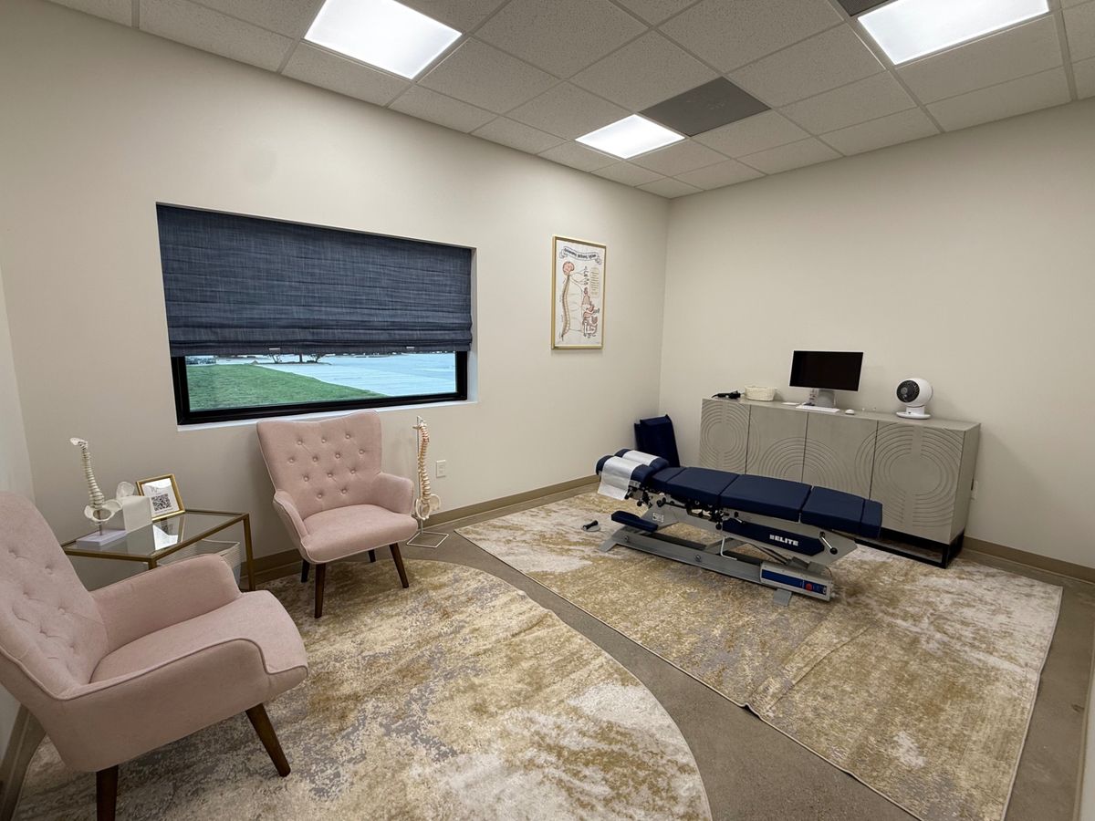 A bright Vitality treatment room with blush armchairs, a patterned rug and an adjusting table